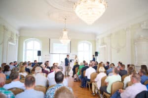 Ein historischer Saal mit Kronleuchtern und Stuckelemeneten. Es sitzen Kommunalvertreter*innen aus Mecklenburg-Vorpommern und hören einen Vortrag auf dem Landeskommunalnetzwerk in Vietgest, ©LEKA MV, Foto: Celien Graubaum