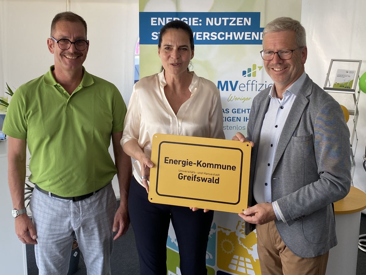 Drei Personen lächeln in die Kamera und halten vor sich eine Plakatte mit der Auszeichnung Energiekommune des Monats. von Links nach rechts Gunnar Wobig- Geschäftsführer der LEKA MV, Staatssekretärin Ines Jesse und Oberbürgermeister der Hansestadt Greifswald Stefan Fassbinder. Im Hintergrund steht ein Banner von MVeffizient.