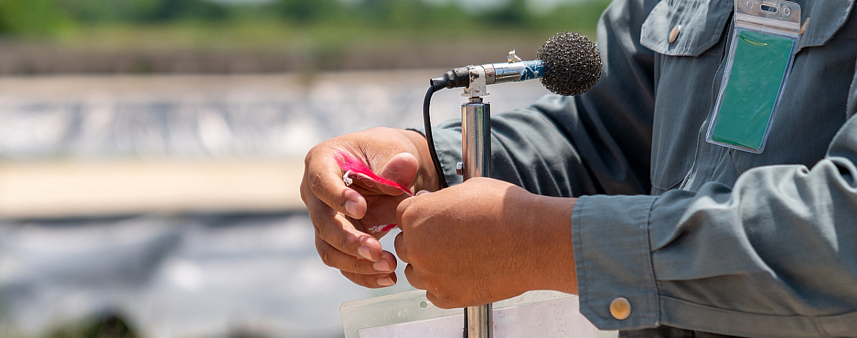 Ein Techniker bereitet eine Schallmessung vor. Er stellt dafür ein Mikrofon in der Landschaft auf und befestigt das Kabel am Stativ, damit keine Nebengeräusche erfasst werden.