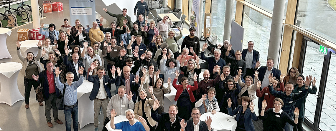 Eine Menschengruppe steht i einem lichtdurchfluteten Foyer und winkt in die Kamera