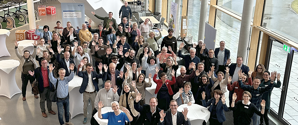 Eine Menschengruppe steht i einem lichtdurchfluteten Foyer und winkt in die Kamera