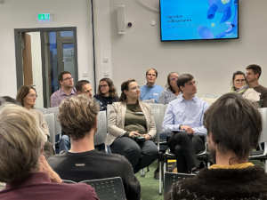 Zuhördende und mitdiskutierende Menschengruppe. Im Hintergrund ist ein großer Bildschirm zu sehen.