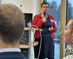 Dr. Beatrix Romberg, Referatsleiterin im Ministerium für Klimaschutz, Landwirtschaft, ländliche Räume und Umwelt Mecklenburg-Vorpommern trägt eine rote Jacke über einem schwarzen Kleid. Sie steht seitlich des Rednerpultes vor einem Terassenfenster. Sie hält ein Mirkrofon in der rechten Hand und hält einen Vortrag. Im Vordergrund sind zwei Zuhörende von hinten zu sehen.