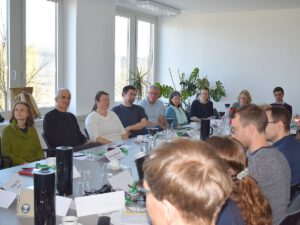 Menschen sitzen an einem Tisch. Im Hintergund ist eine Fentserfront