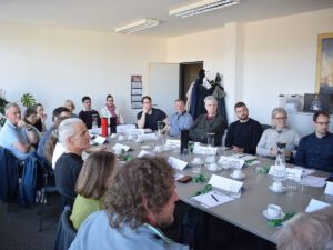 Menschen sitzen in Arbeitsatmophäre um einen Tisch auf dem Getränke stehen und Arbeitsmaterialien liegen.