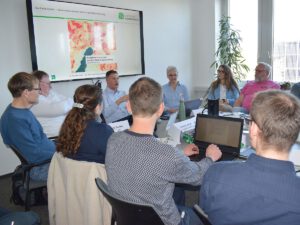 Teilnehmende am Frühjahresentzwerktreffen der Klimaschutzmanager*innen. Sie sitzen an einem Tisch auf dem Arbeitsmaterialien, Laptop und Getränke stehen an der Wind ist eine digitale Leinwand.