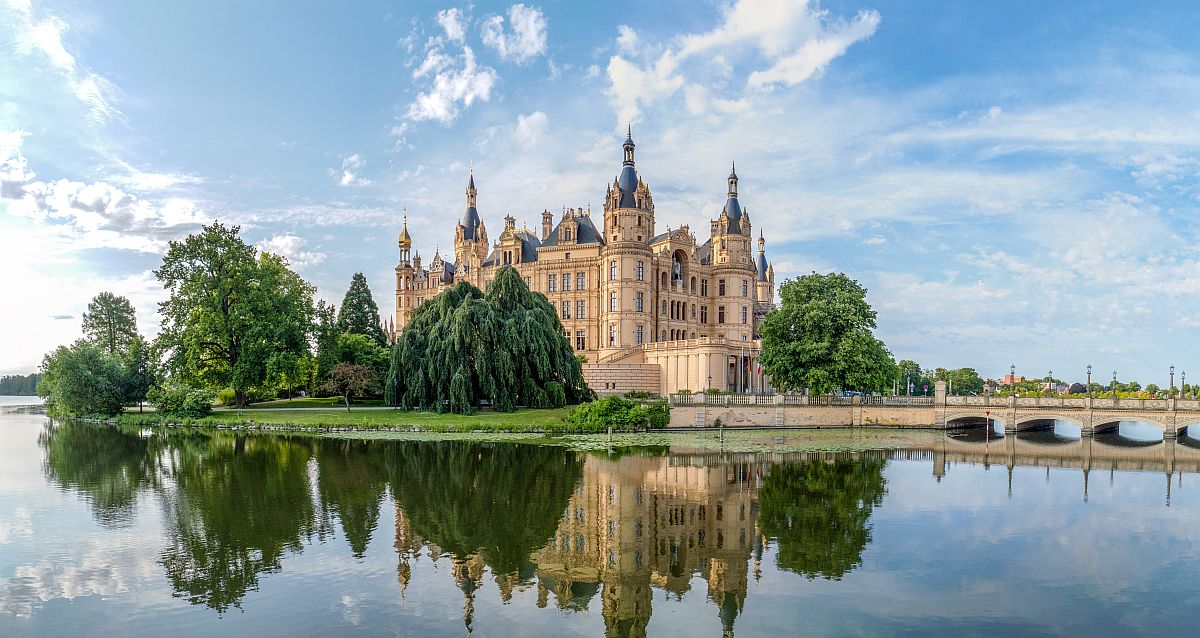 Das Schweriner Schloss ist ein prachtvolles, märchenhaftes Bauwerk. Es hat viele Türme, vergoldete Kuppeln und eine sehr prunkvolle Fassade. Das Schloss liegt malerisch auf einer Insel im Schweriner See und ist über eine Brücke zu erreichen. Um das Schloss befinden sich stattliche begrünte Bäume. Sie Szene spiegelt sich im Wasser.
