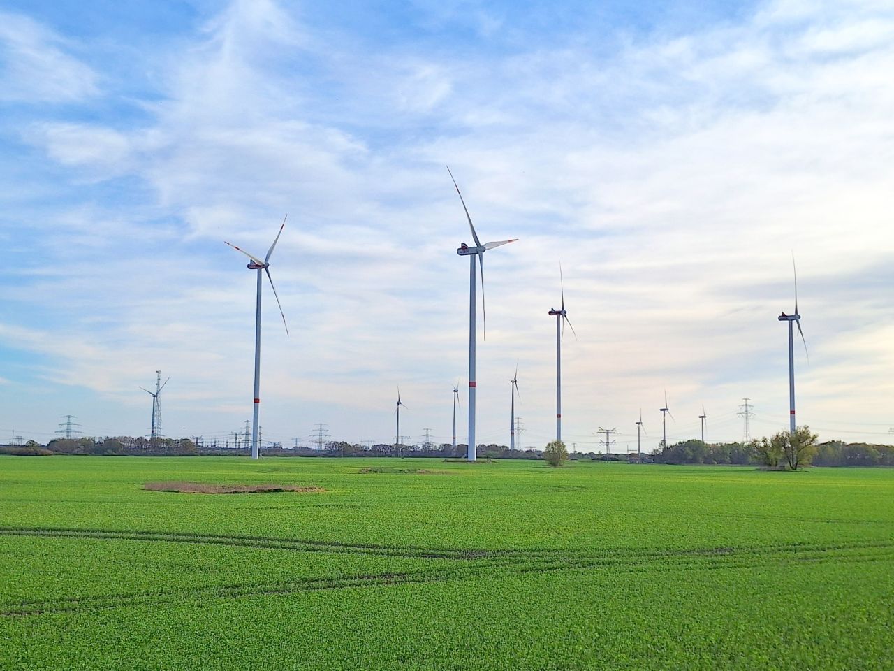 Ein Windpark auf grünem Acker vor bewölkten Himmel Die Rotoren sind nach rechts im Bild ausgerichtet die Flügelspitzen sind rot.