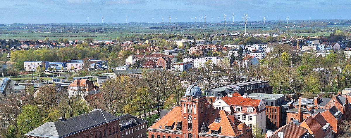 Luftaufnahme der Hansestadt Greifswald. Am Horizont sind Winenergieanlagen zu sehen. Im Vordergrund ist das hitorische Physikalische Institut mit Sternwarte. Es ist ein altes Backsteingebäude.