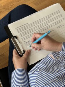 Jemand schreibt etwas mit blauen Stift auf ein Blatt Papier, was auf dem Klemmbrett geheftet ist.